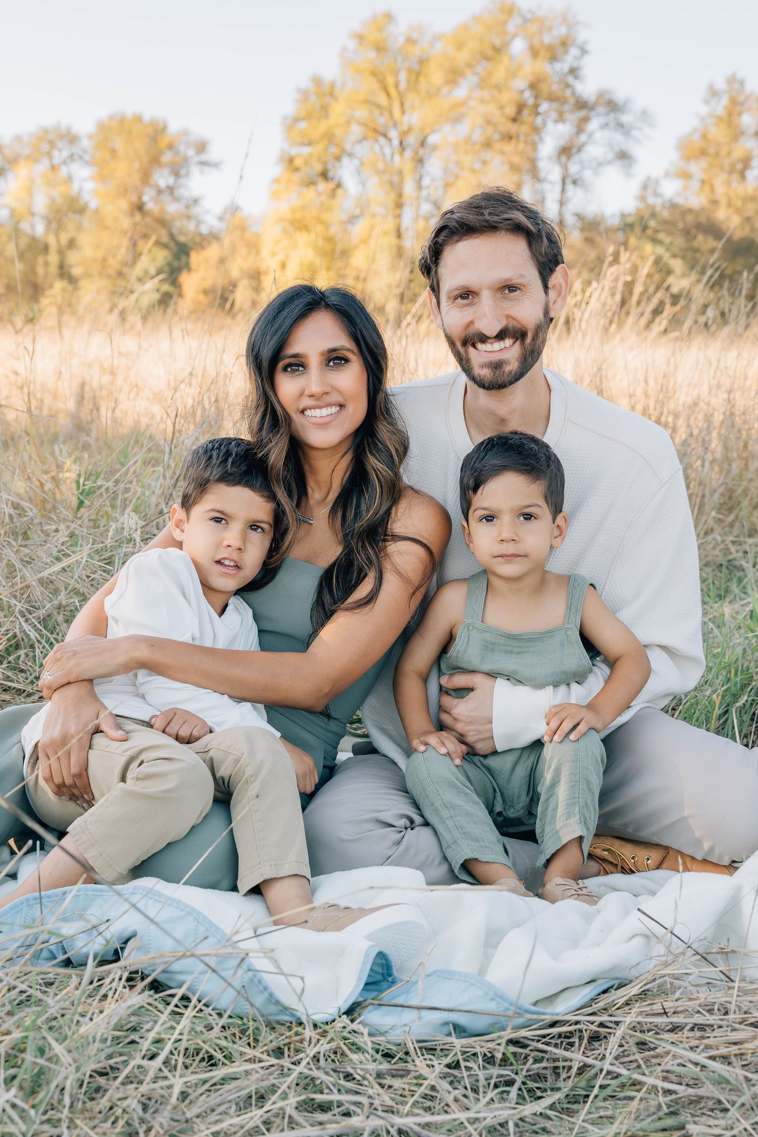 Joyful family of four sitting together on a blanket in a golden field during a warm fall photo session.