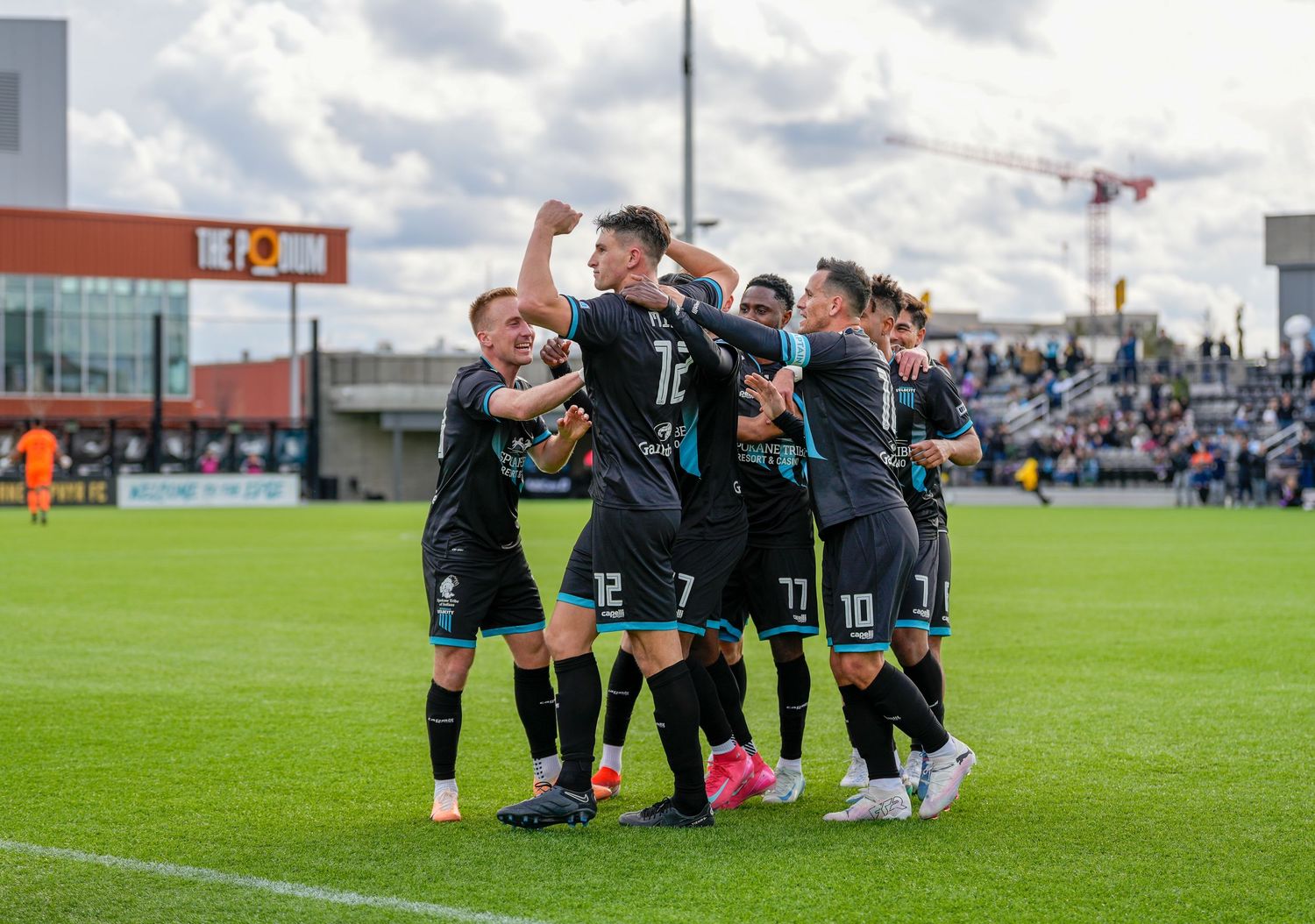 Soccer players celebrate together on the field during a match at a stadium with spectators in the background.
