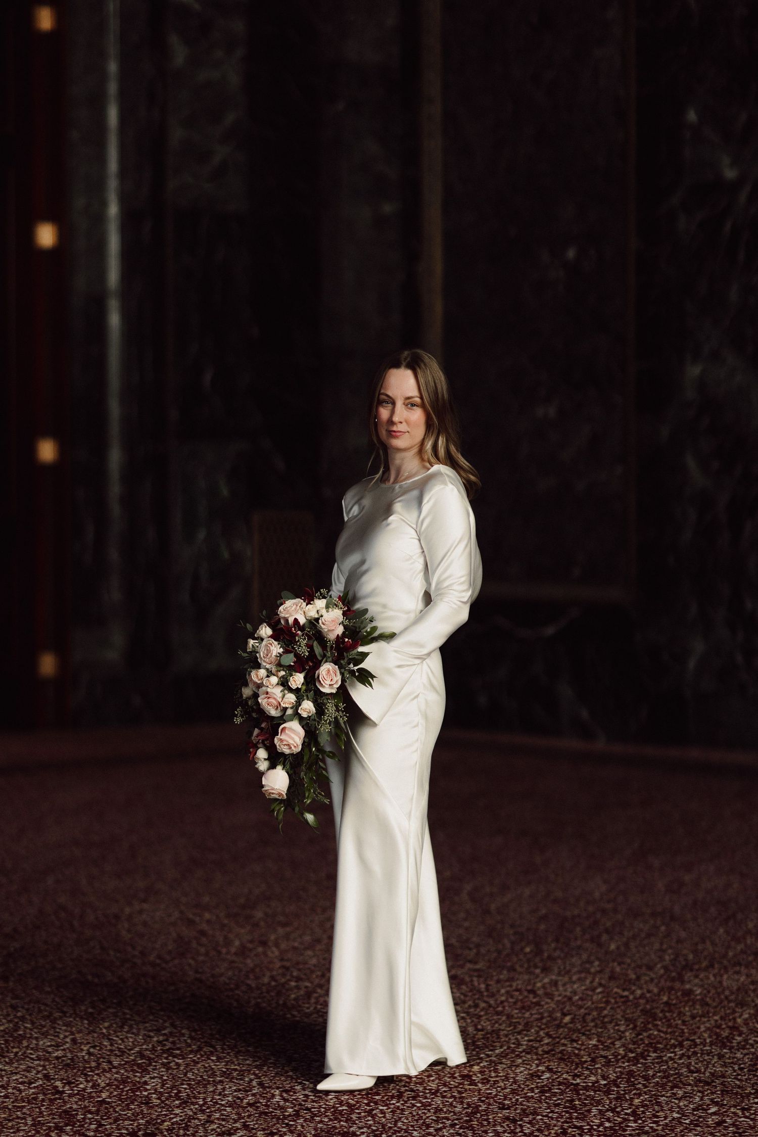 Bride in elegant white long-sleeved dress holds romantic bouquet in dark moody setting.
