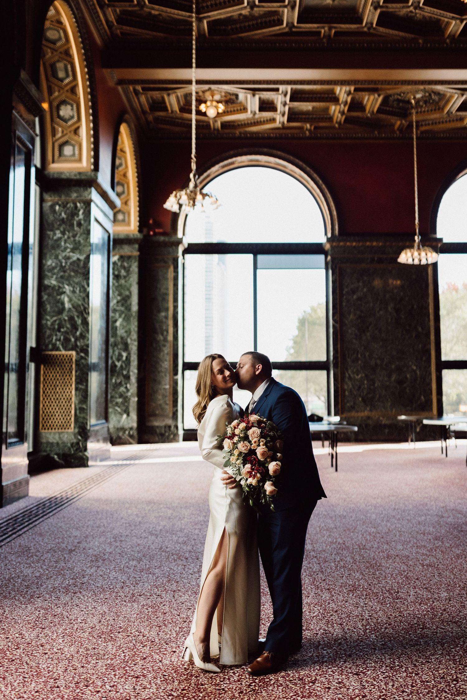 Couple embraces in grand historic hall with ornate architecture and arched windows.