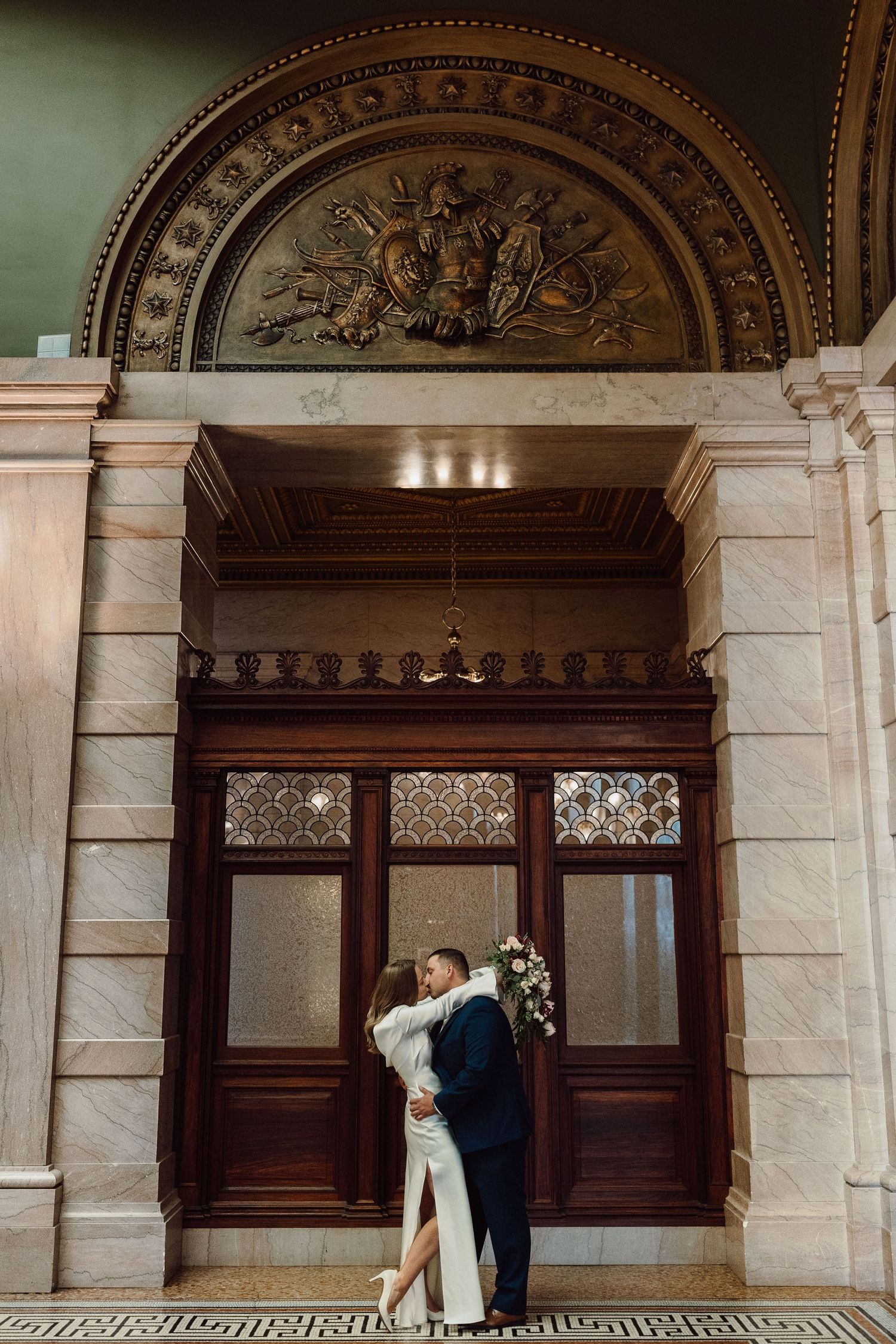 Wedding couple shares intimate moment in classical marble hallway with decorative arches.