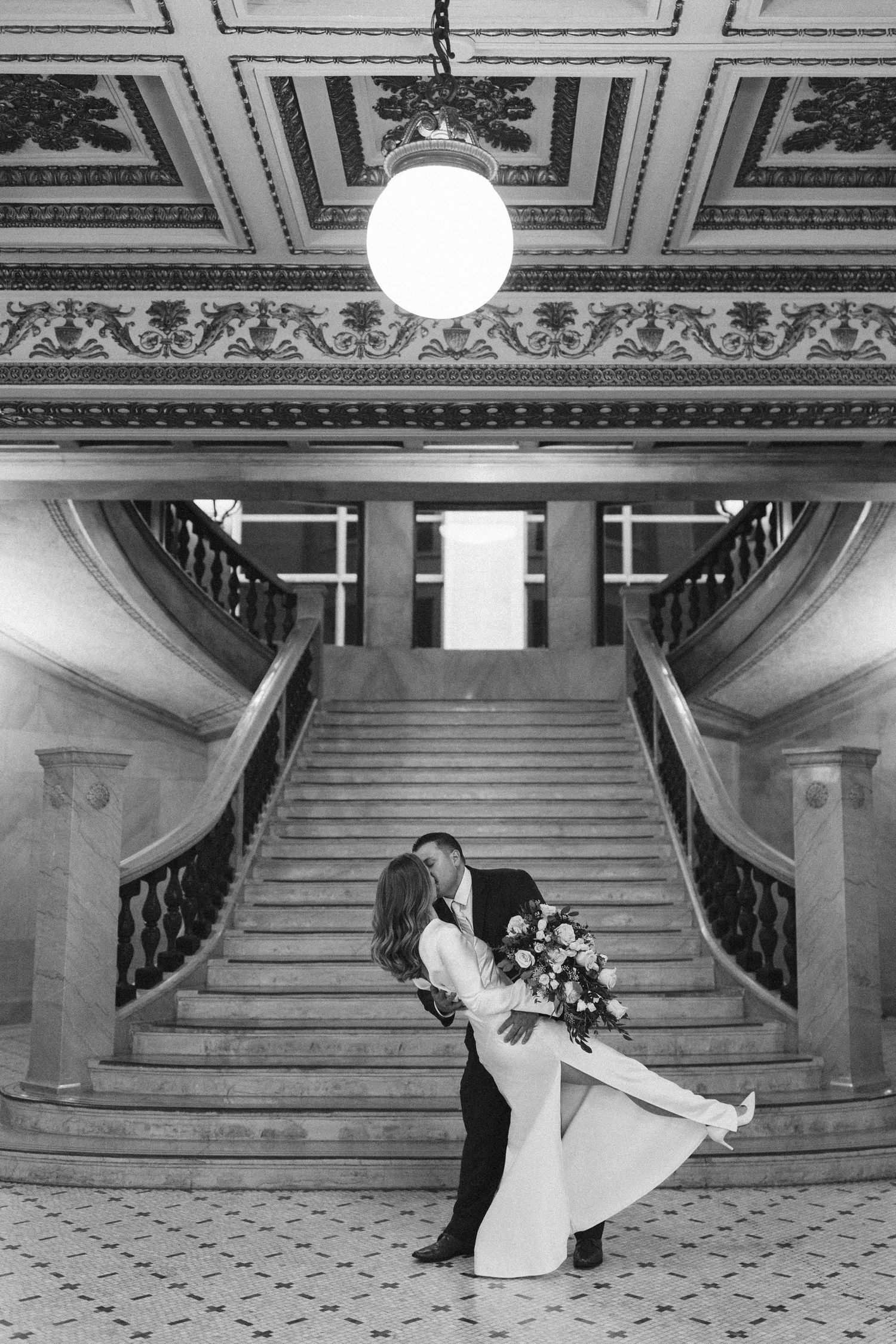 Black and white photo of couple dancing in grand hallway with ornate ceiling and staircase.