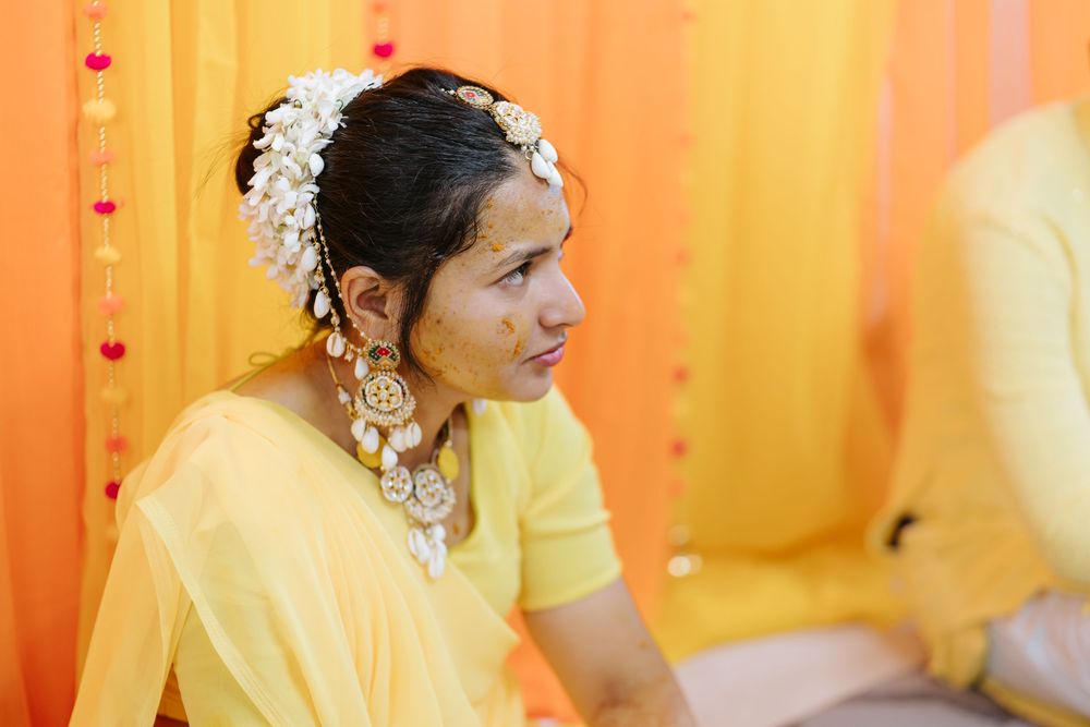 A person in a yellow traditional outfit with floral hair accessories and jewelry sits against an orange backdrop.