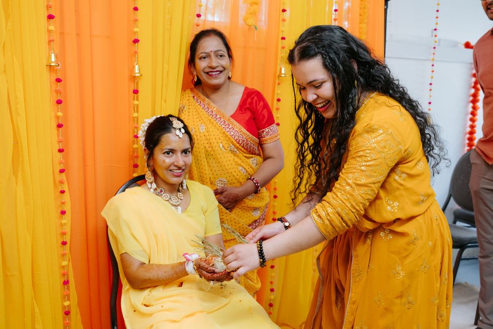 Traditional Indian haldi ceremony with people wearing vibrant yellow and red outfits against orange backdrop with decorative lights.