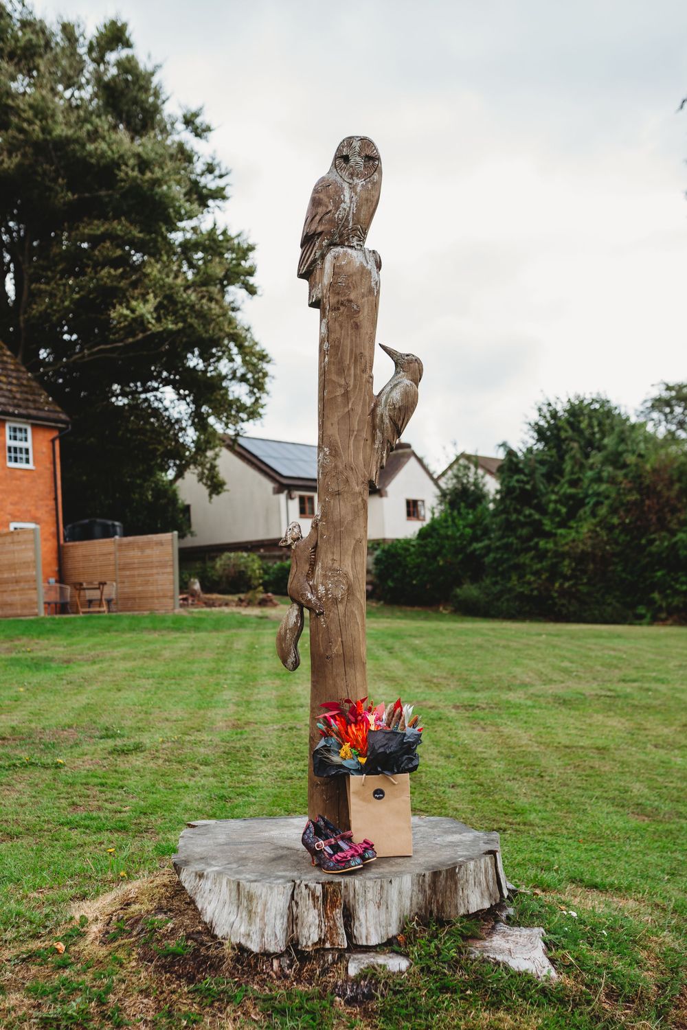 Carved wooden post with owls perched on top stands in a garden with houses in the background.