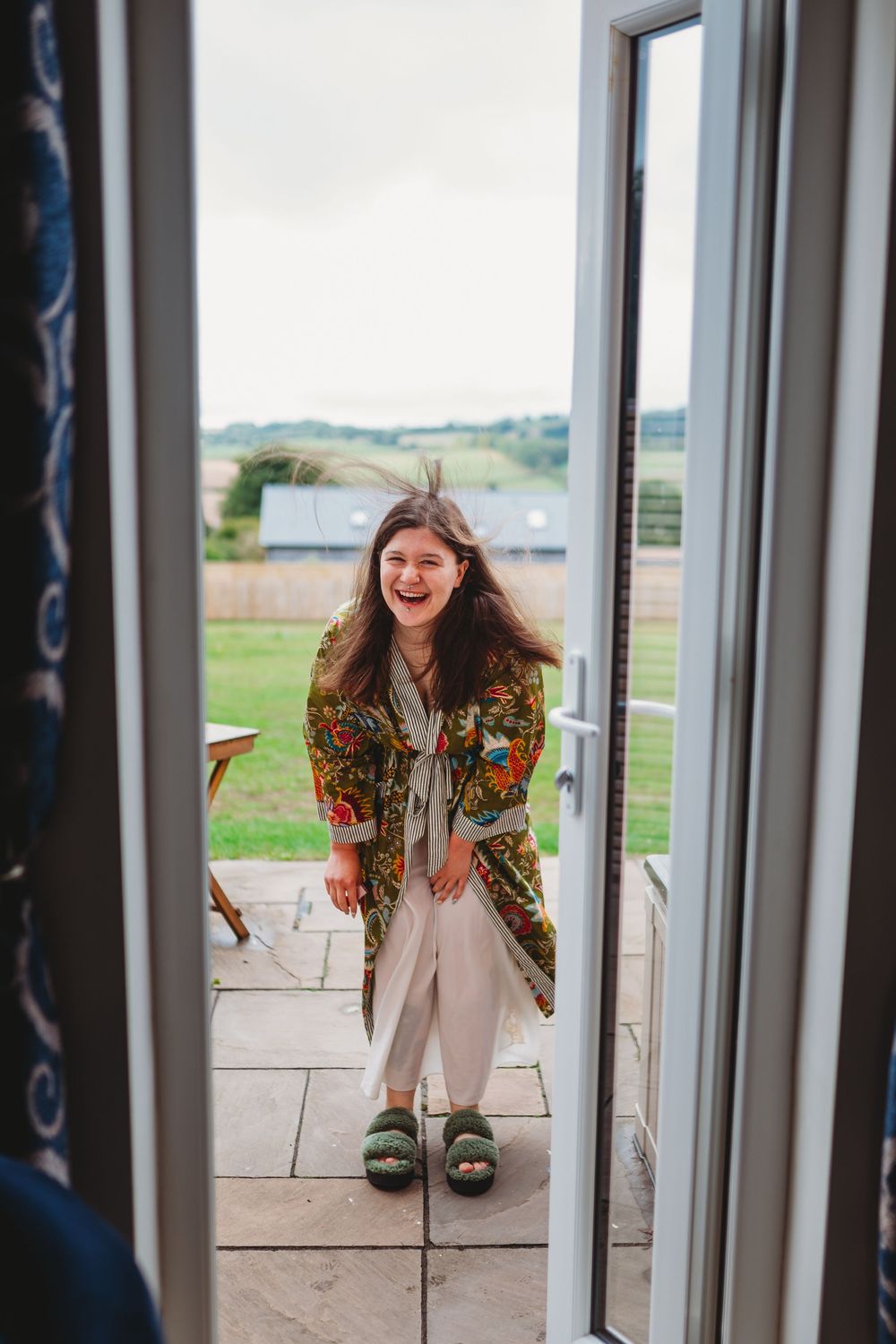 Person wearing a floral robe stands laughing in a doorway with countryside views behind them.