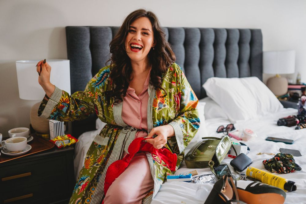Person in colorful floral robe sits joyfully on bed during wedding morning preparations.