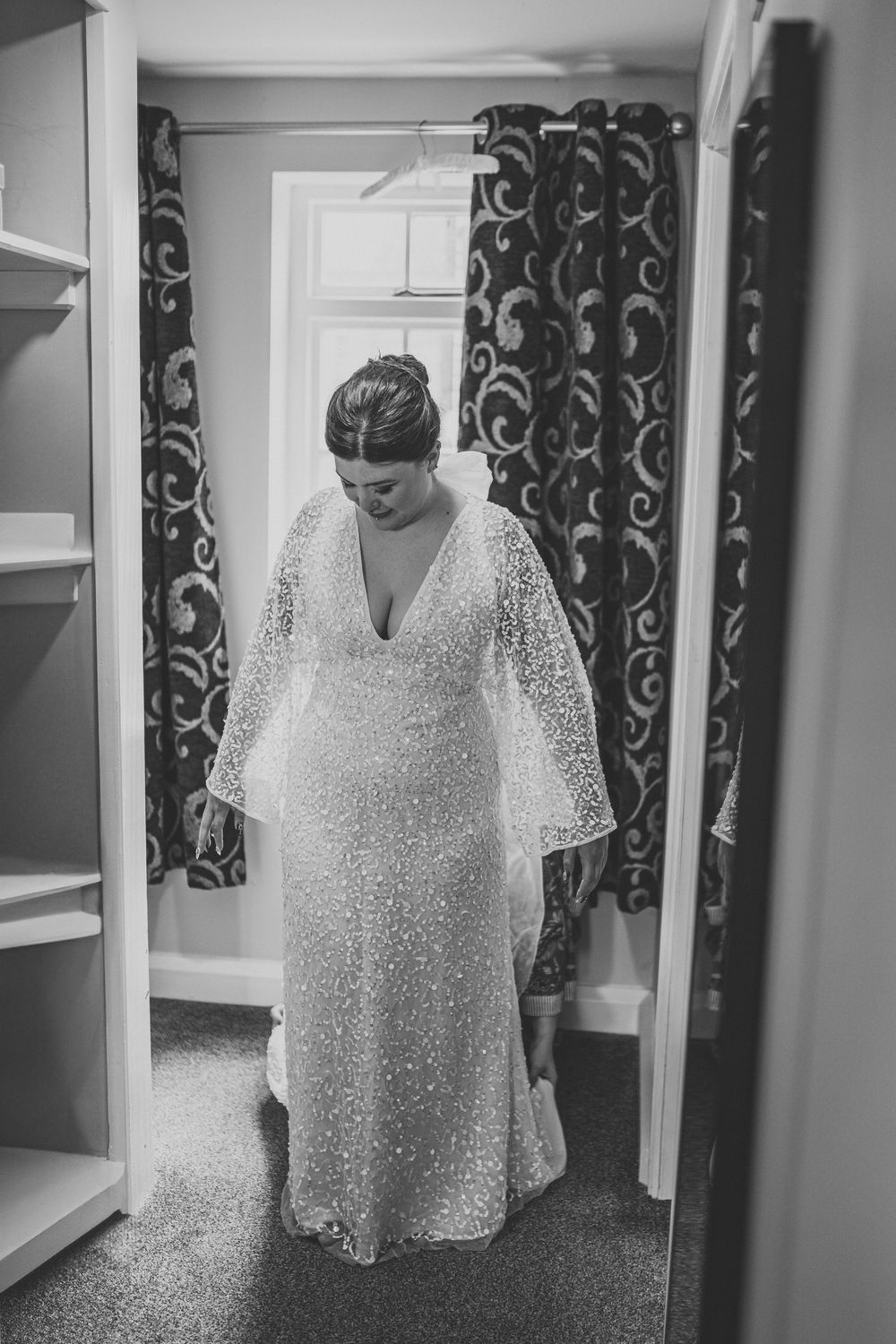 Person wearing white lace wedding gown stands in dressing room with patterned curtains.