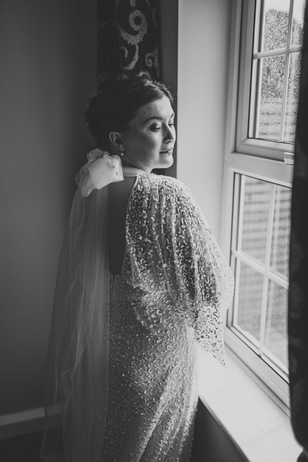 Black and white portrait of a wedding dress with intricate beading by a window.