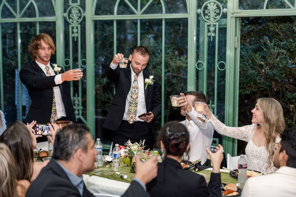 Wedding guests raise champagne glasses in celebration at an elegant greenhouse venue during a reception.