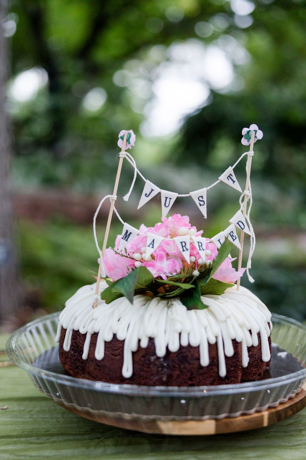 A chocolate cake with white dripping icing and pink flowers on top sits on a plate in an outdoor garden setting.