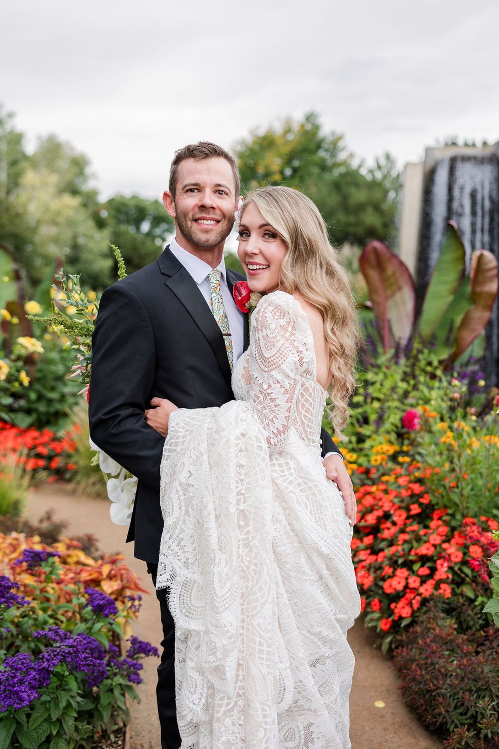 A couple poses together in a vibrant garden filled with orange and purple flowers on their wedding day.