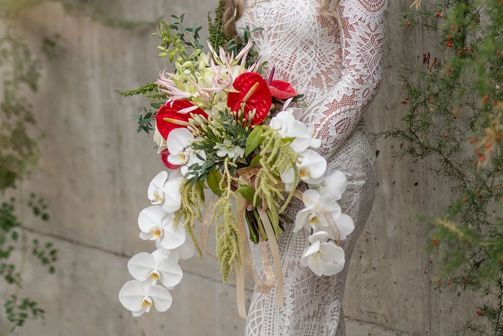Elegant white lace wedding dress detail with cascading white orchids and red floral bouquet accents against stone wall.