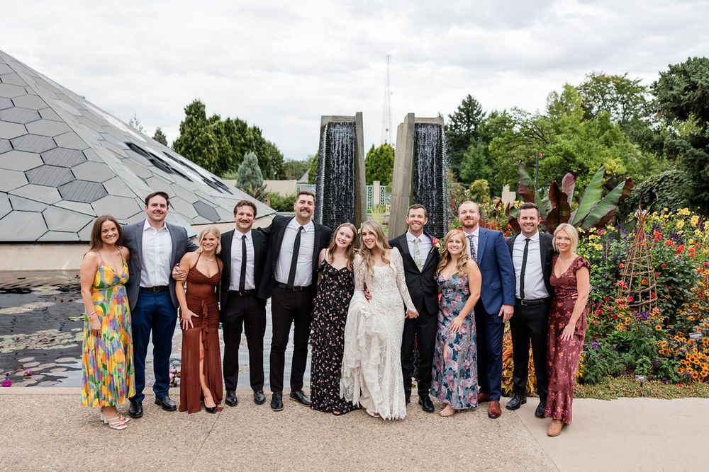 A large wedding party group photo taken outside against a modern architectural backdrop.