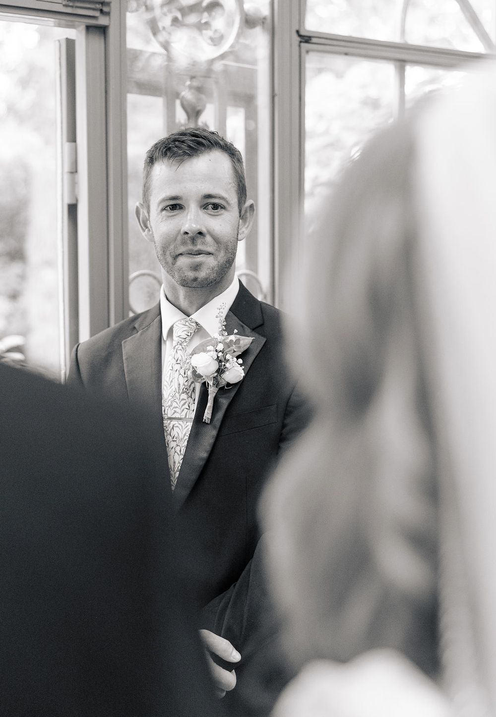 Black and white wedding photo showing groom in formal suit with boutonniere and patterned tie at indoor ceremony.