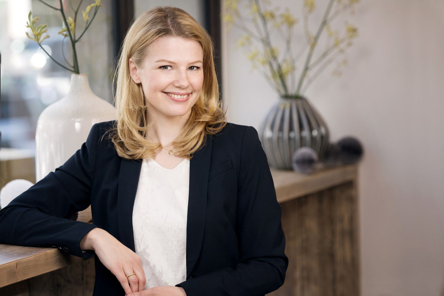Professional woman in black blazer and white blouse poses confidently in modern office setting.