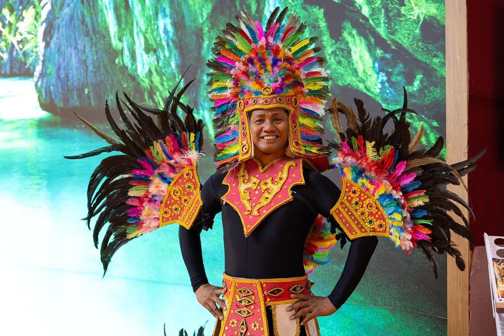 Colorful carnival dancer wearing vibrant feathered headdress and costume performs against turquoise backdrop at the WTM in the ExCel, London