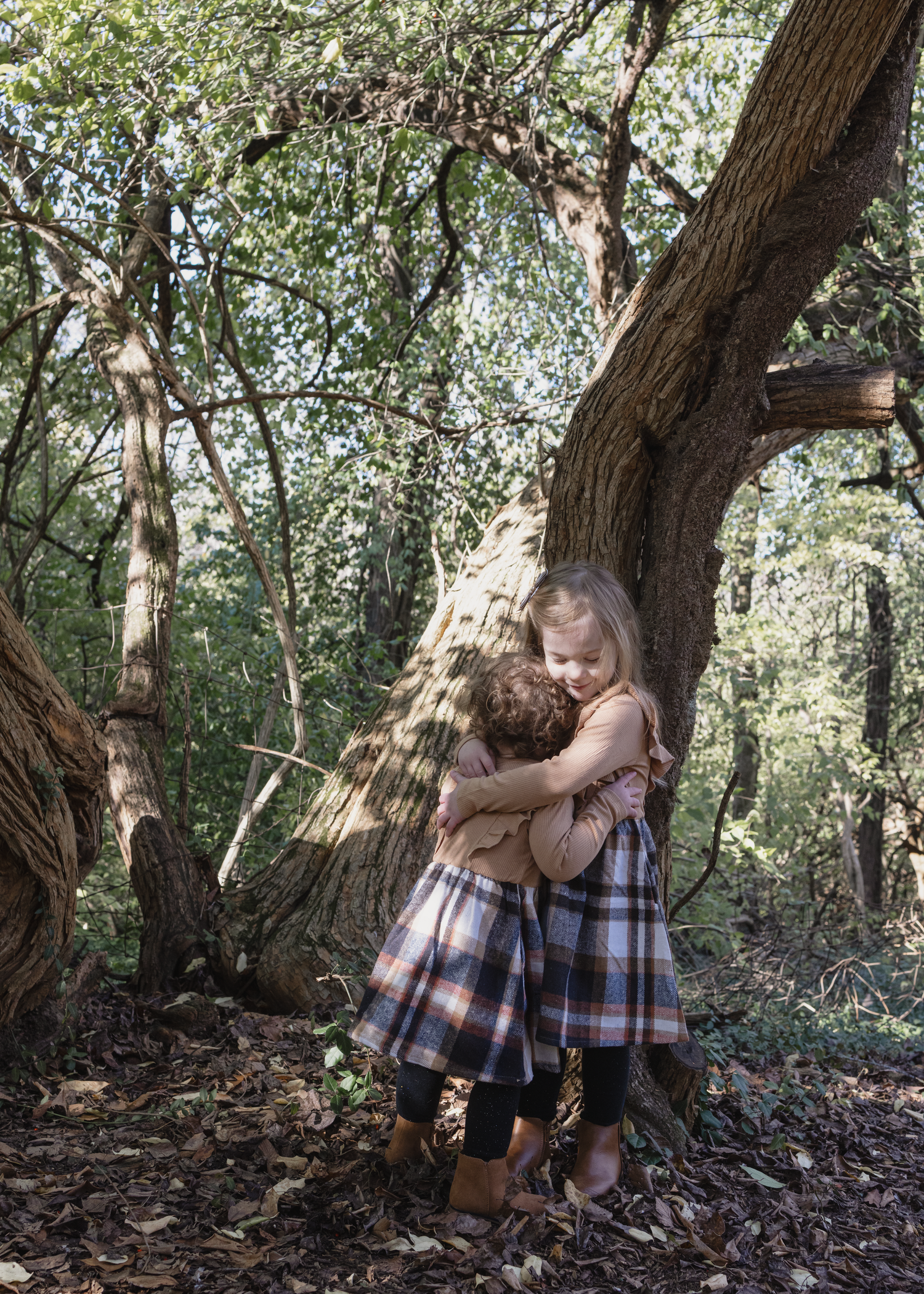 Two small children share a tender hug while standing near a large tree trunk in a wooded forest setting.