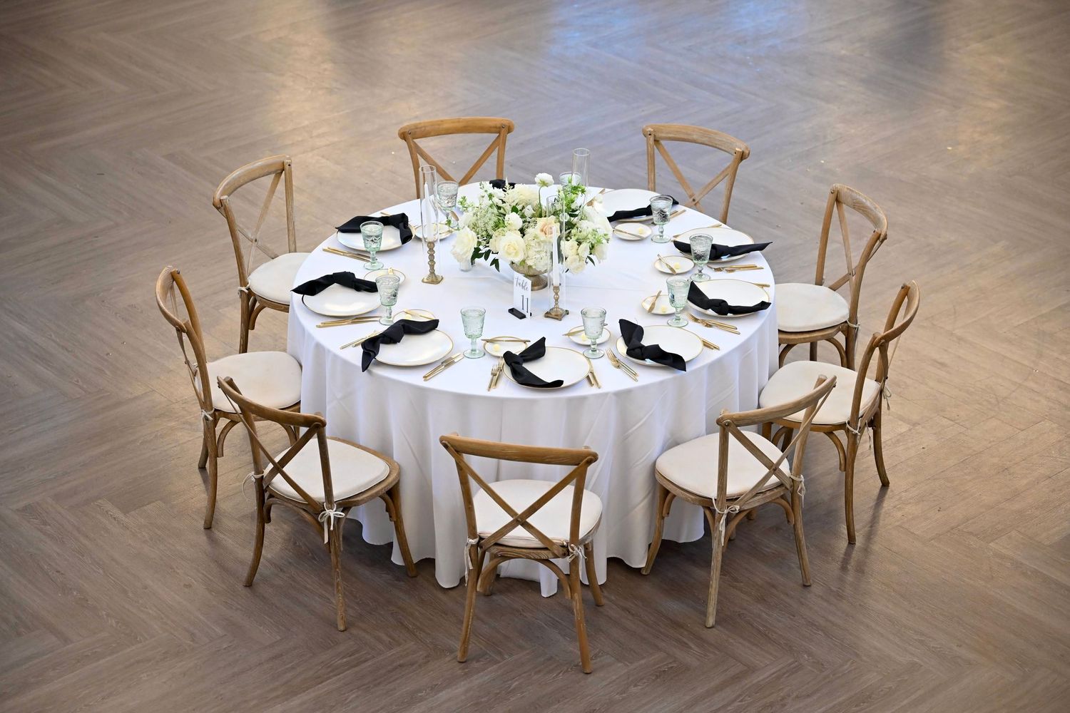 Round white dining tables with wooden crossback chairs arranged in a banquet hall setting on polished hardwood floors.