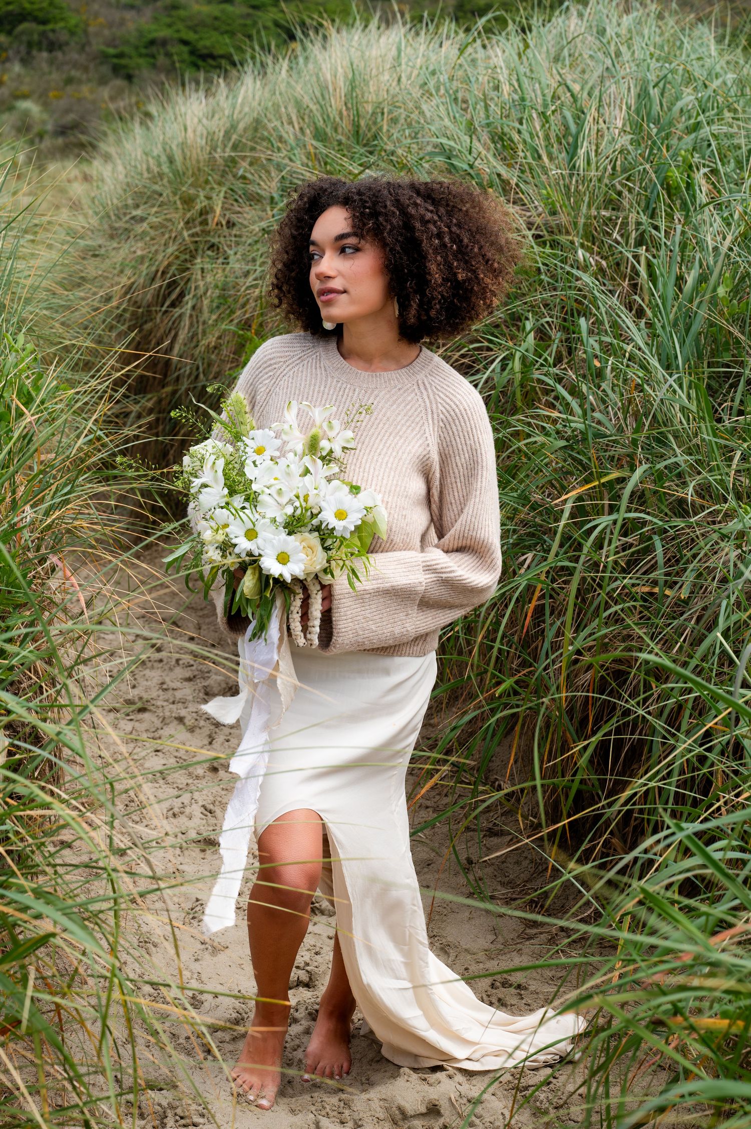 A bride in a cream sweater and dress poses with white flowers among tall beach grass on a sandy dune.