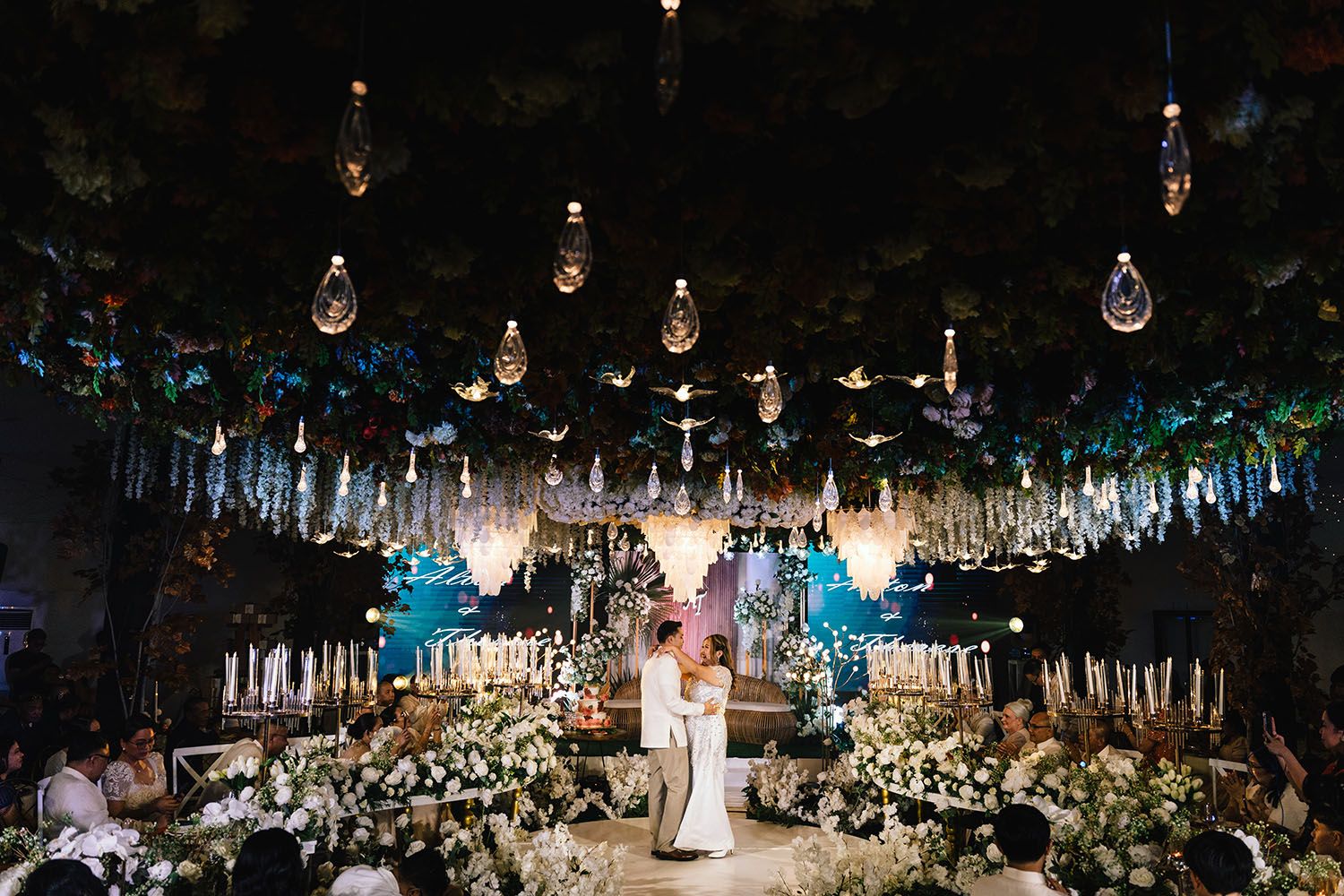 Hanging light bulbs create a magical atmosphere at an outdoor evening wedding reception with white floral arrangements.