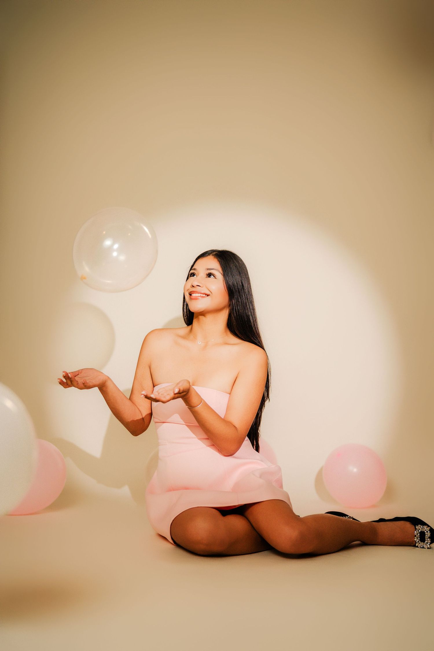 A person in a pink dress poses playfully with floating balloons against a beige backdrop in a studio setting.