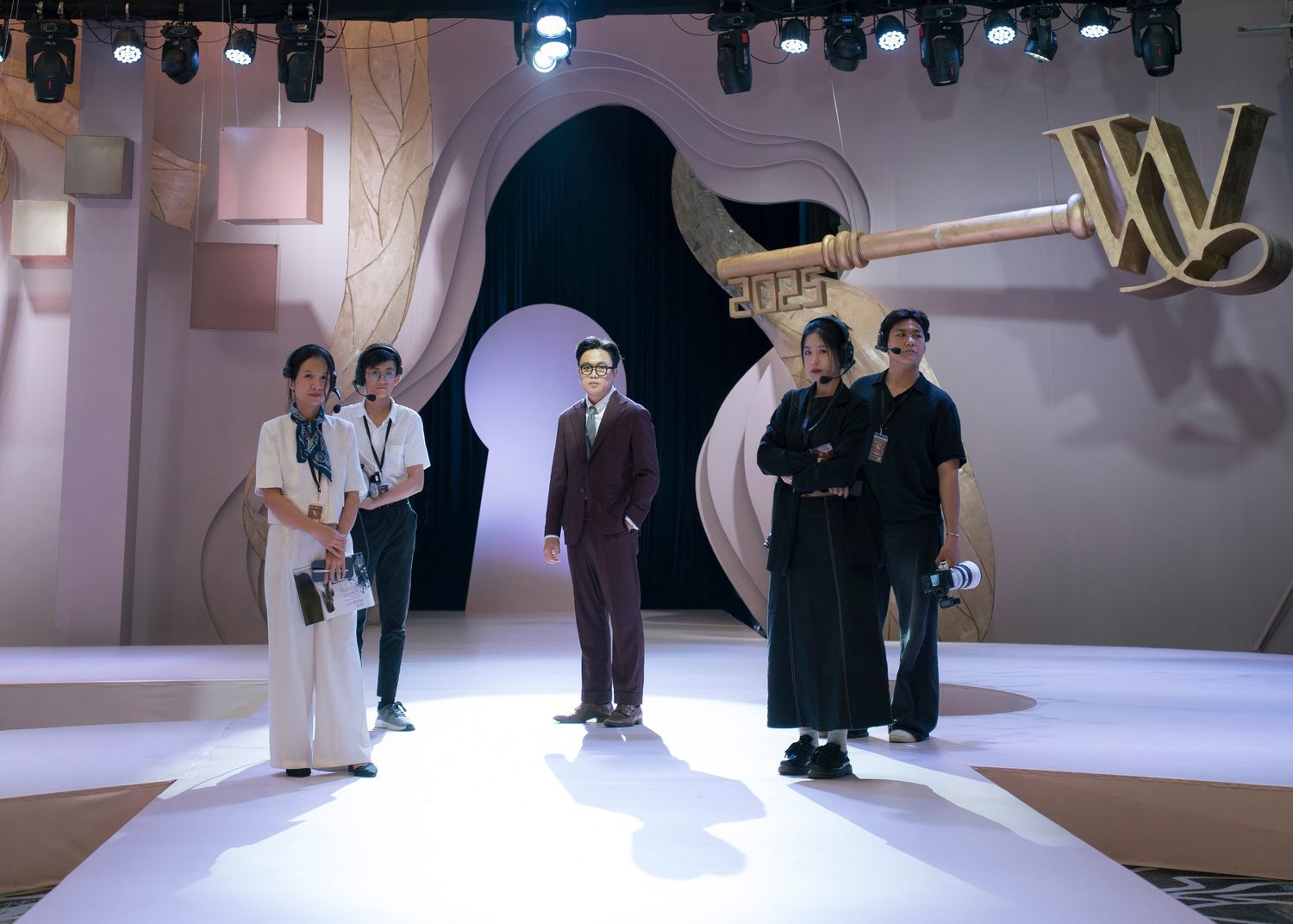 Group of five people posing on stage runway beside large decorative key backdrop at fashion event.