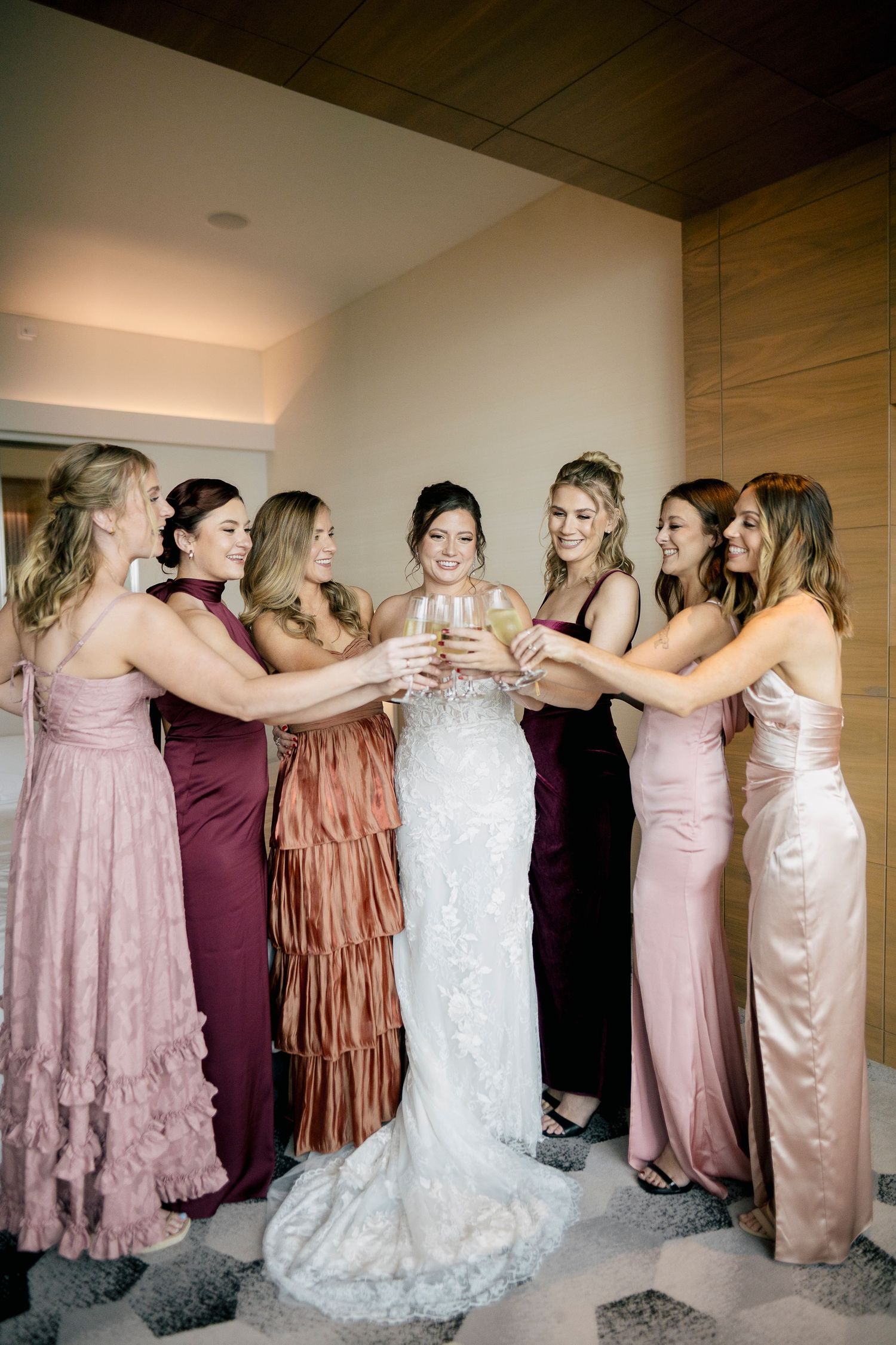 A line of bridesmaids in pink and burgundy dresses embrace each other in a conga line formation at a wedding.