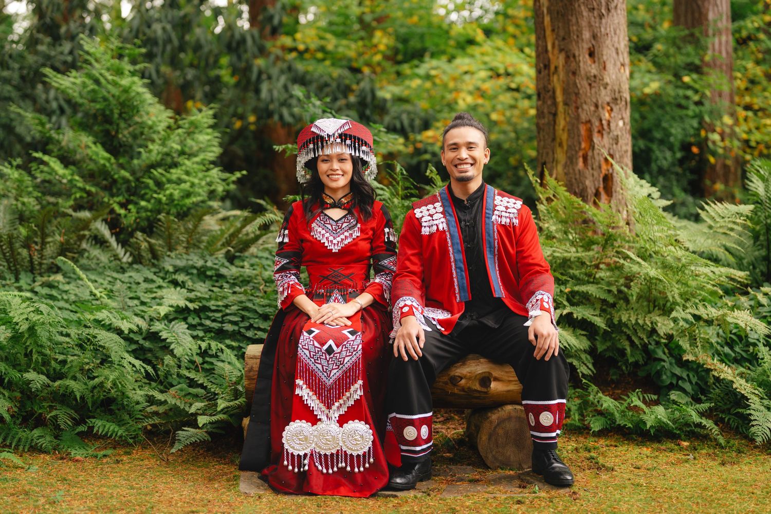 A couple in traditional Norwegian folk costumes sit on a wooden bench surrounded by lush green ferns in a forest setting.