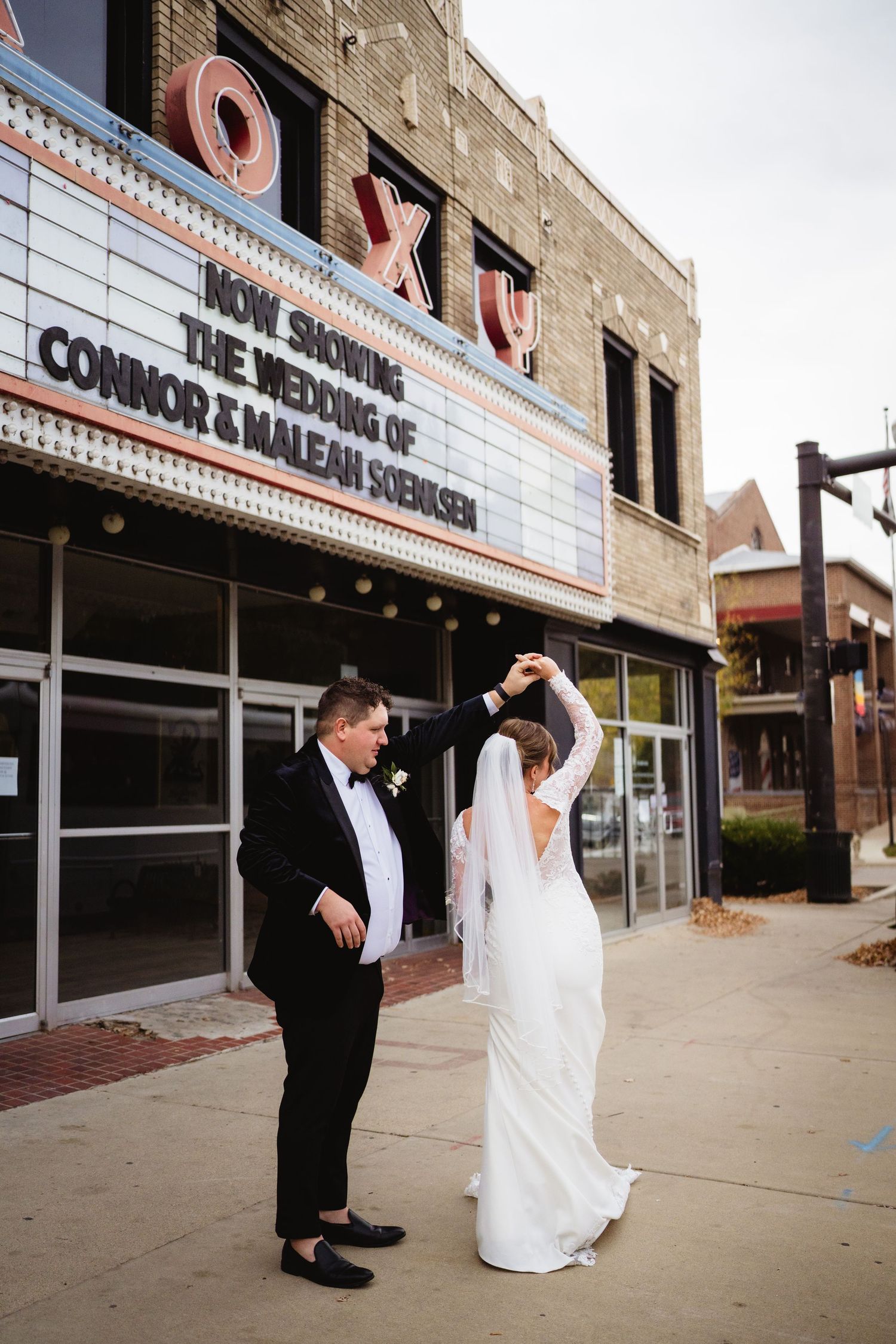 A couple in wedding attire dances playfully outside a vintage movie theater with a retro marquee sign.