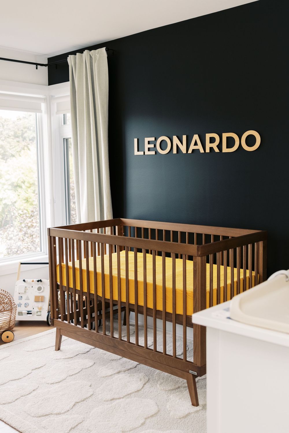 Stylish navy blue nursery featuring walnut crib with mustard bedding and gold LEONARDO letters above near window.