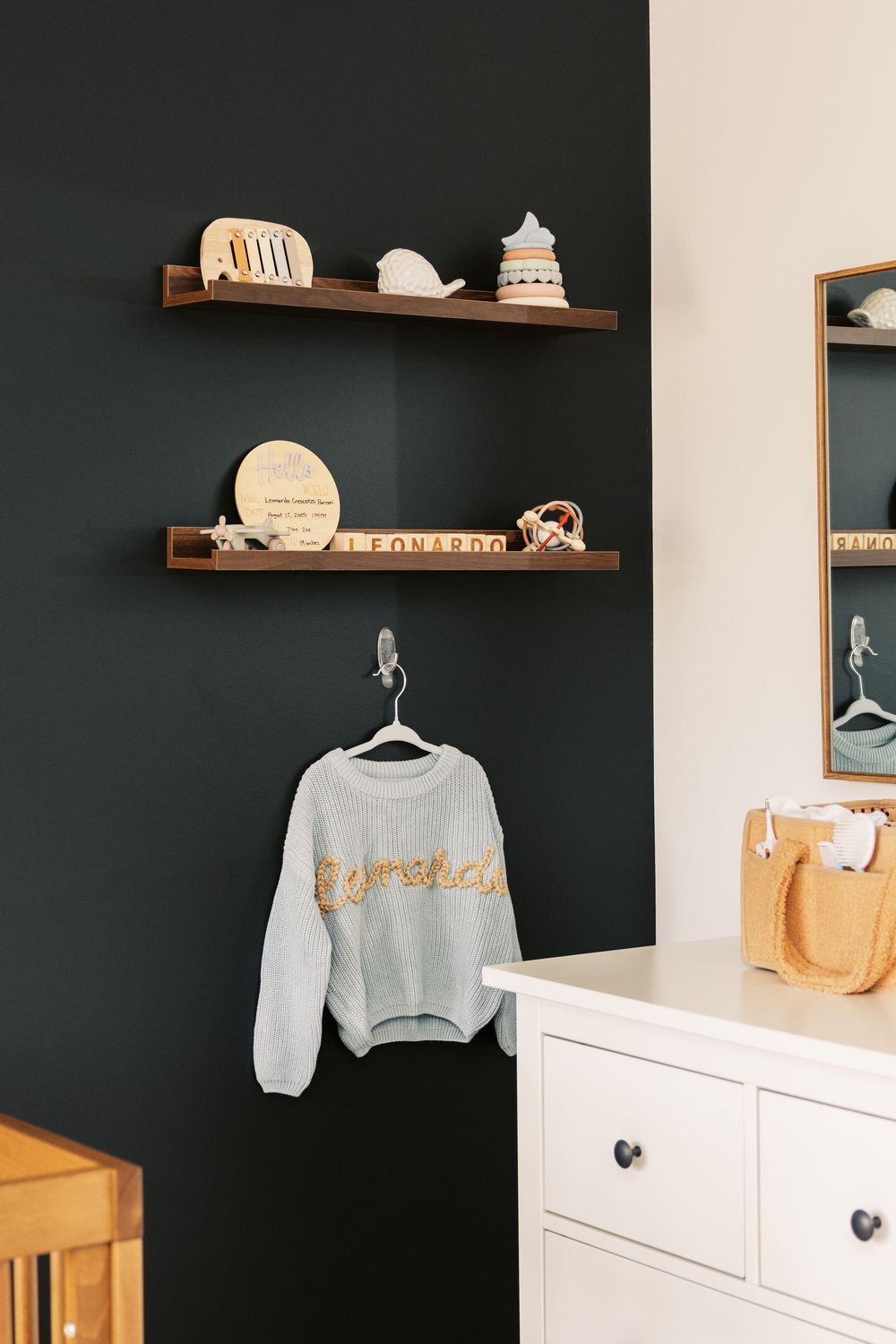 Dark nursery wall with floating walnut shelves displaying baby keepsakes and personalized Leonardo knit sweater below.