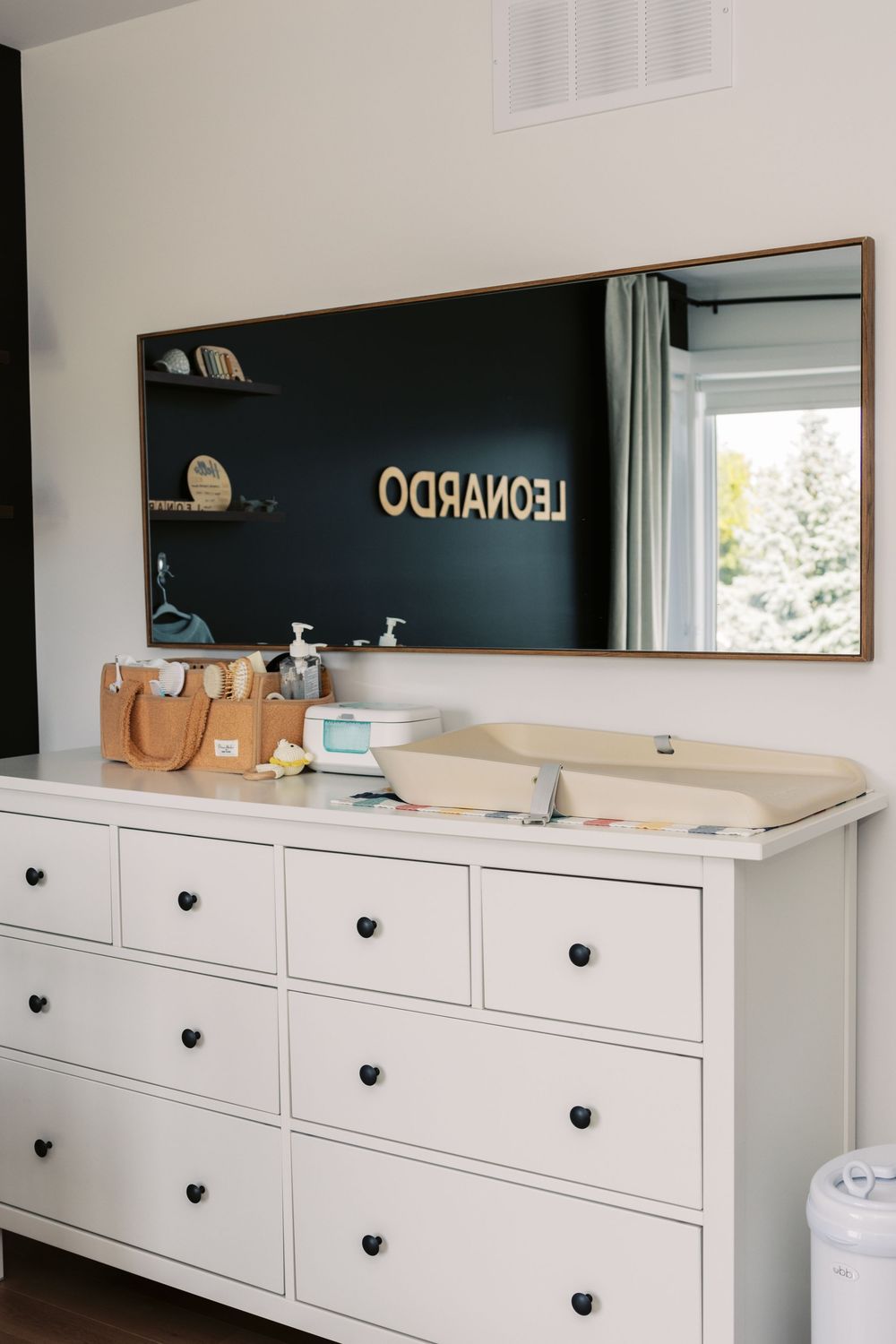White dresser with changing pad topped with a large mirror reflecting the name LEONARDO on a dark nursery wall.