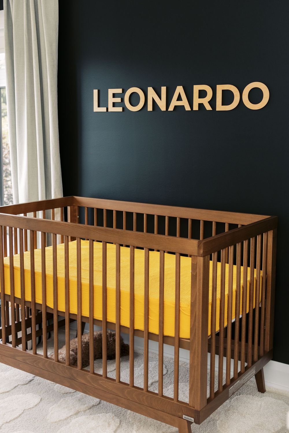Modern navy nursery with walnut wood crib, mustard yellow sheet, and gold LEONARDO name letters on dark accent wall.