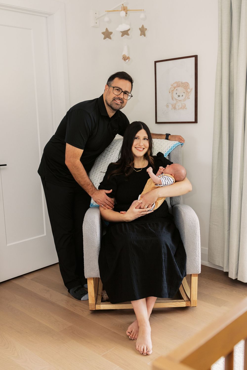 New parents in matching black outfits pose with newborn baby in a cozy nursery with star mobile and lion wall art.