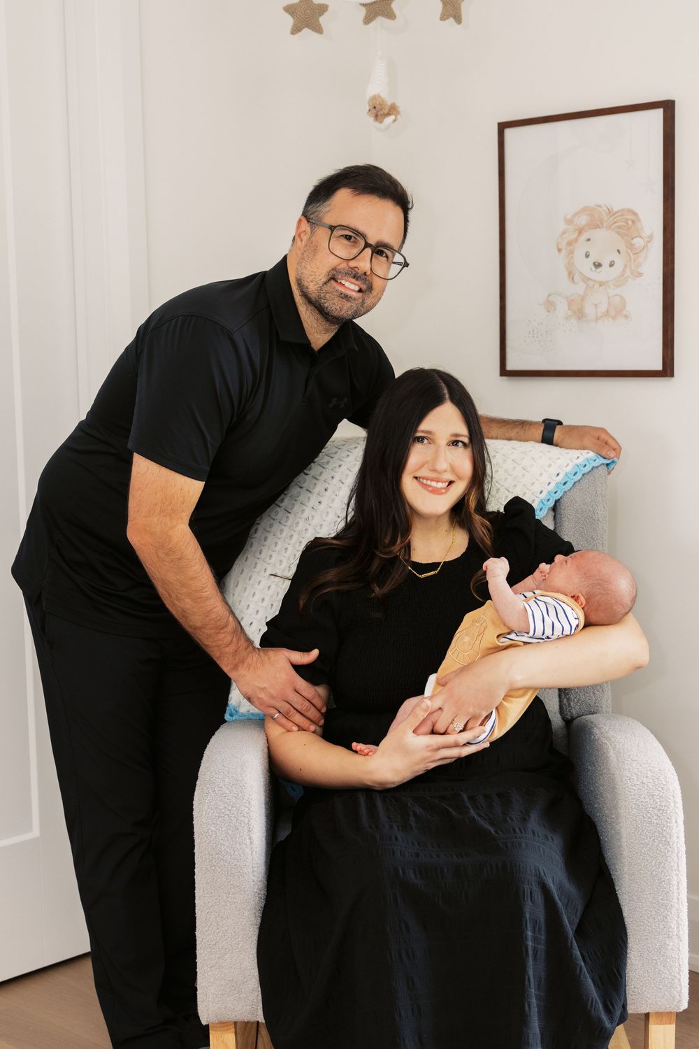 Happy new family of three with newborn baby poses in gray nursery rocking chair beneath star mobile and lion art.