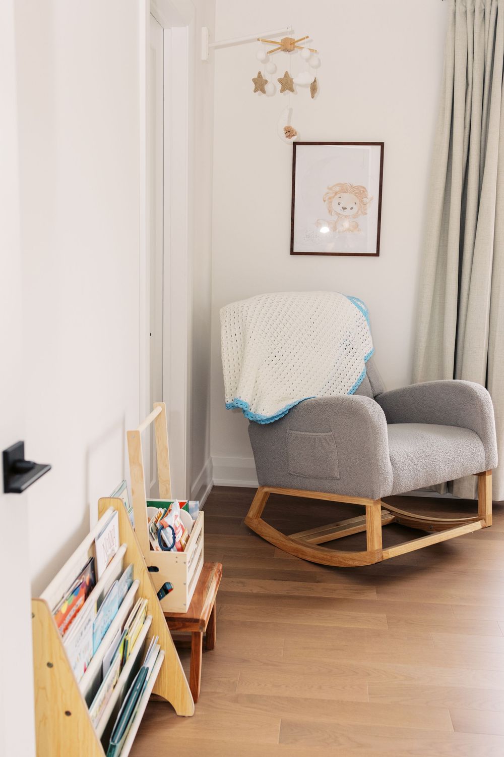 Cozy nursery corner with gray rocking chair, knit blanket, wooden bookshelf, star mobile, and lion wall art print.