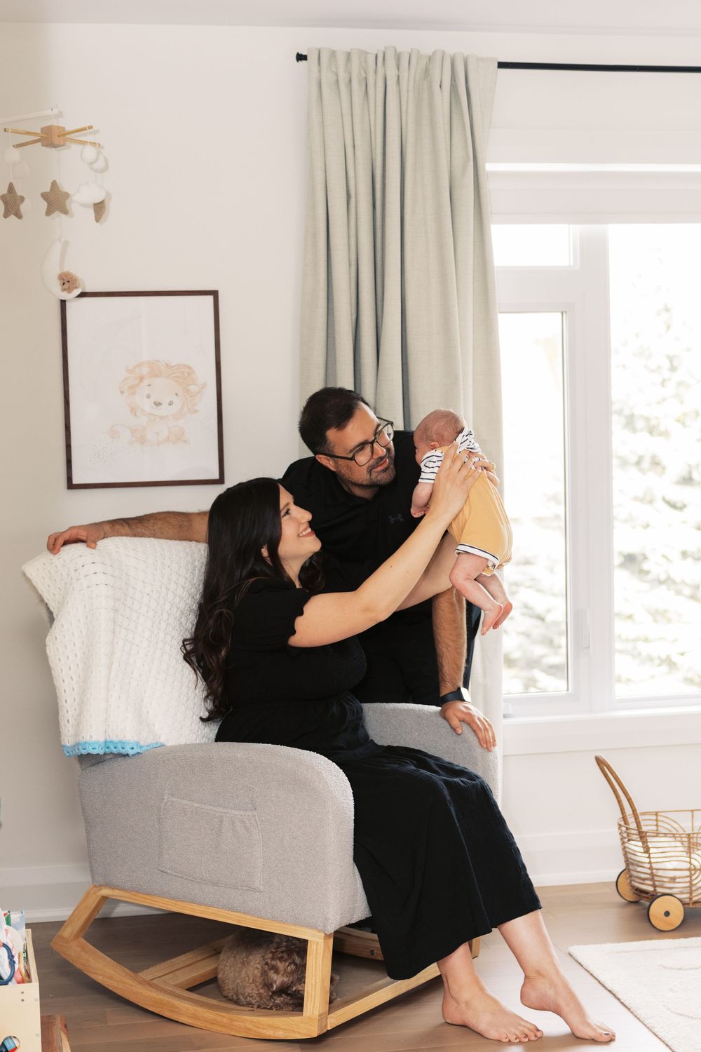 Happy couple in black outfits admire their newborn baby in a bright nursery with rocking chair and lion artwork.