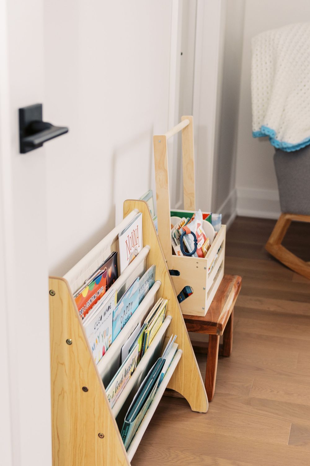 Wooden Montessori-style bookshelf displaying colorful childrens books beside a small step stool in a bright nursery.