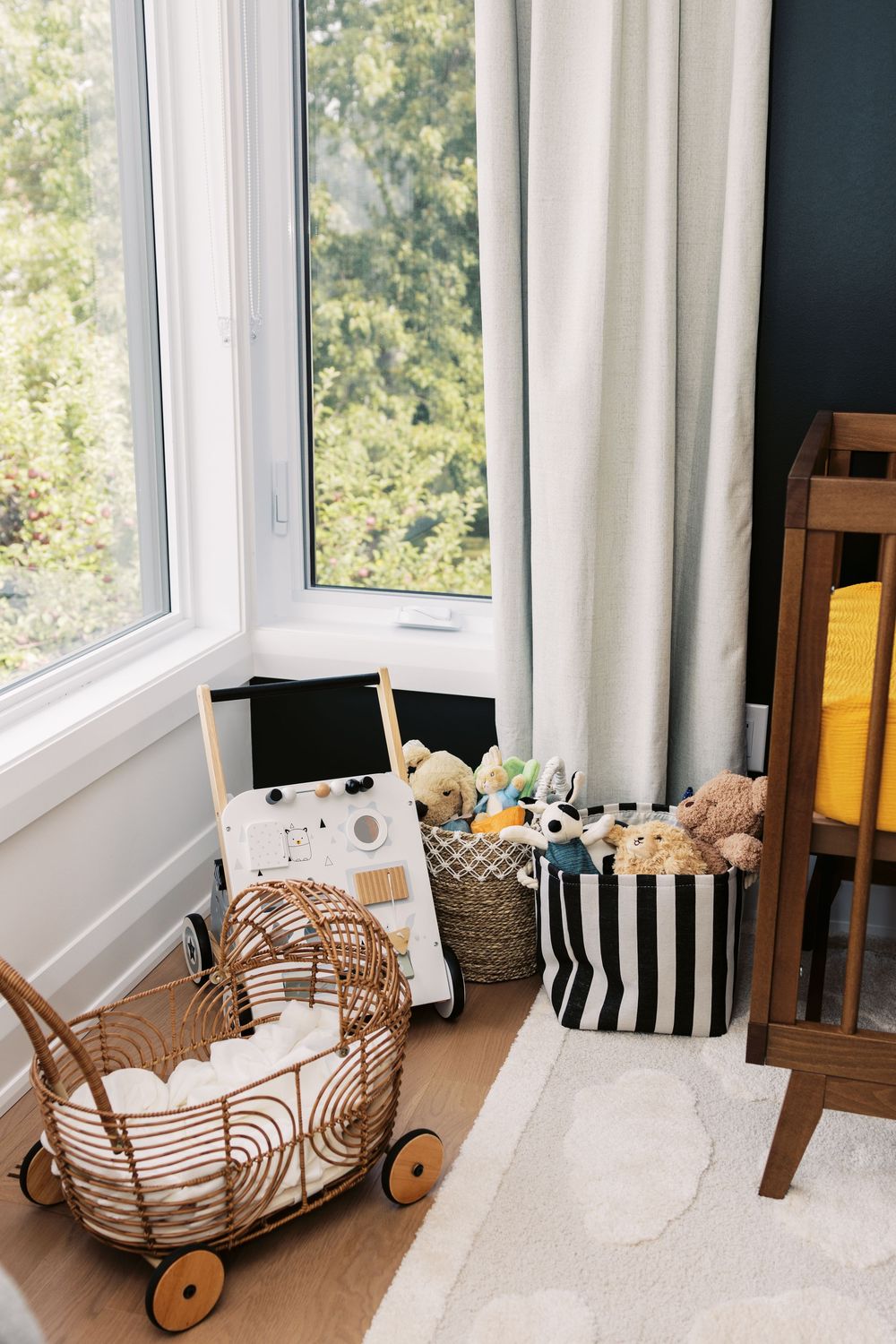 Cozy nursery corner with rattan doll pram, stuffed animal baskets, and white curtains beside a bright window.