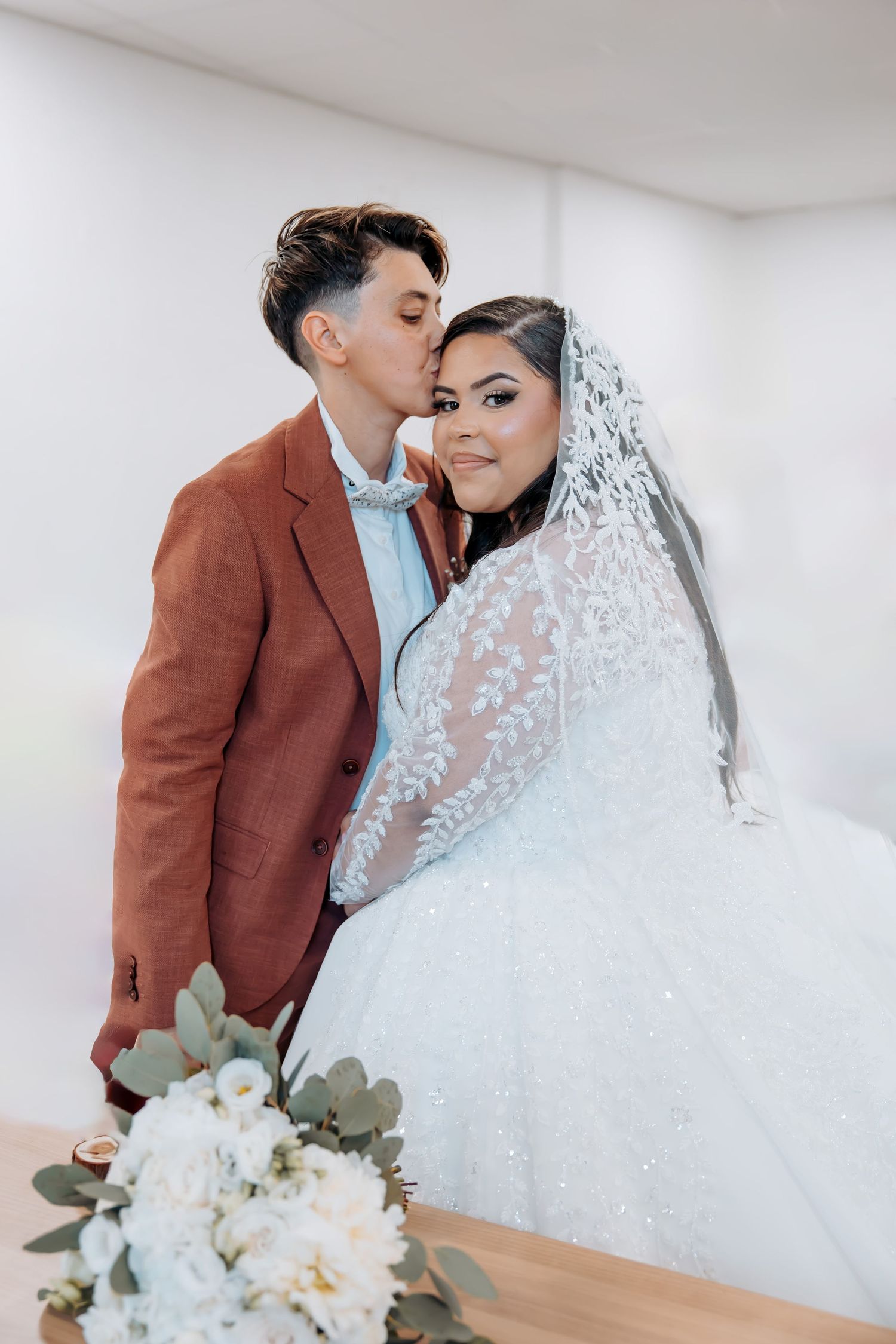 Intimate wedding portrait with couple in white dress and brown suit against white wall.