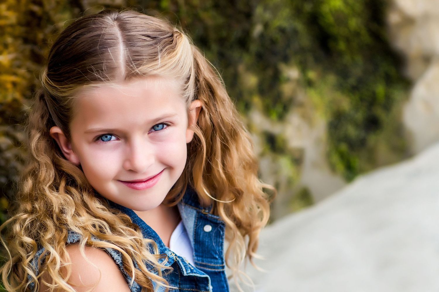 Young person with curly blonde hair wearing denim vest outdoors.