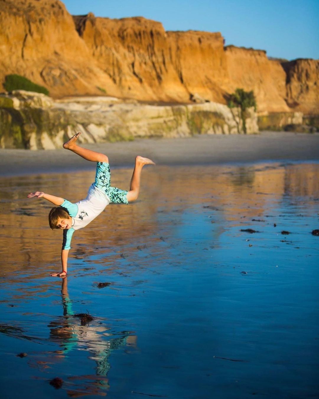 Person doing a one-handed handstand on wet beach sand with cliffs in background.