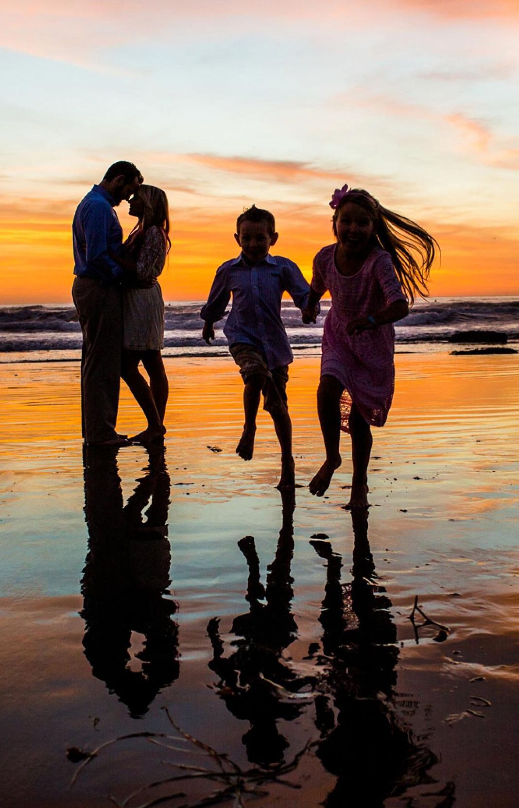 Silhouettes of family playing and running on beach at sunset with reflections in wet sand.
