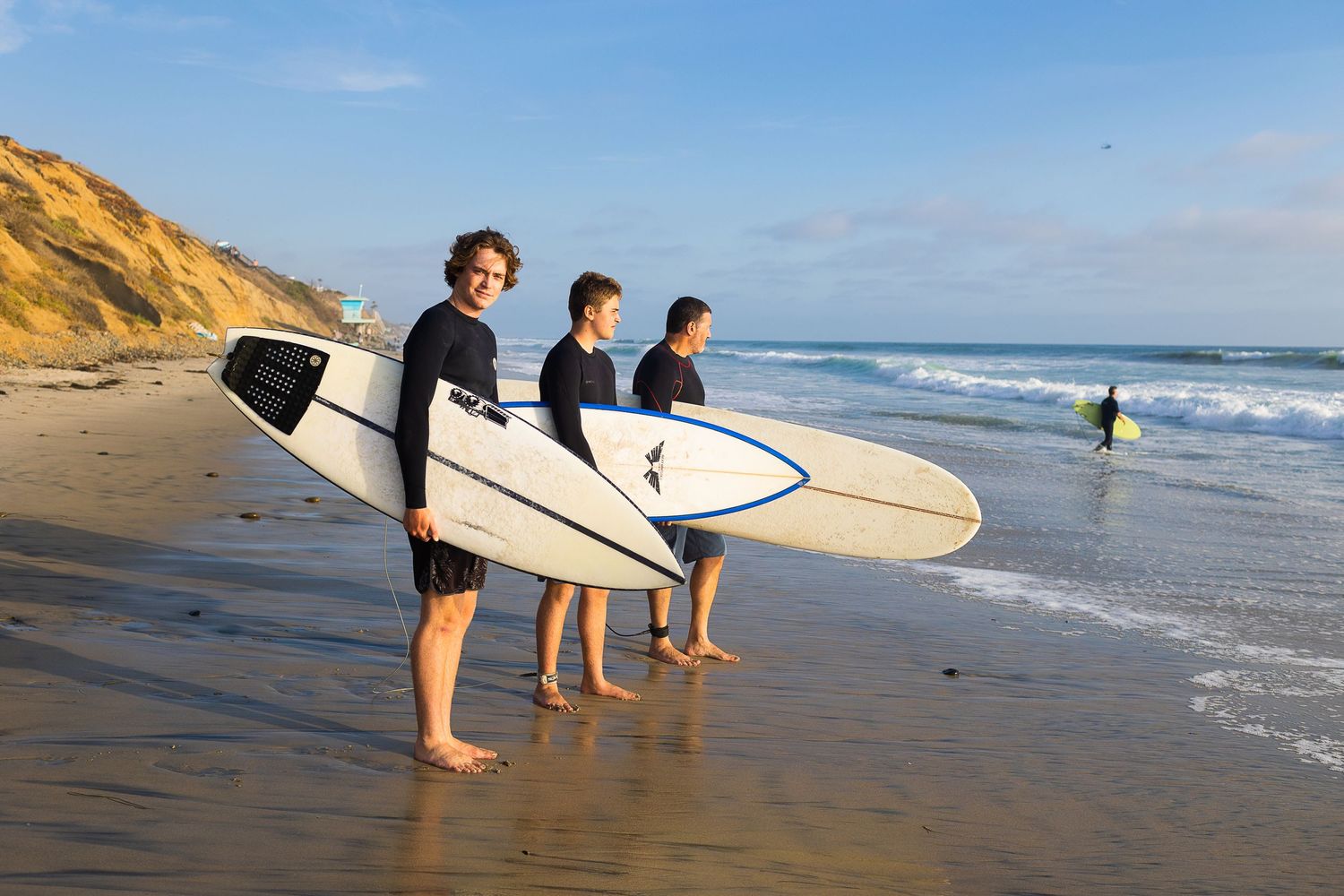 Three surfers carrying surfboards along a sunny beach with cliffs in the background.