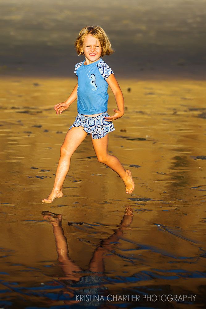 Child jumping on wet sand beach at sunset creating mirror reflection below.