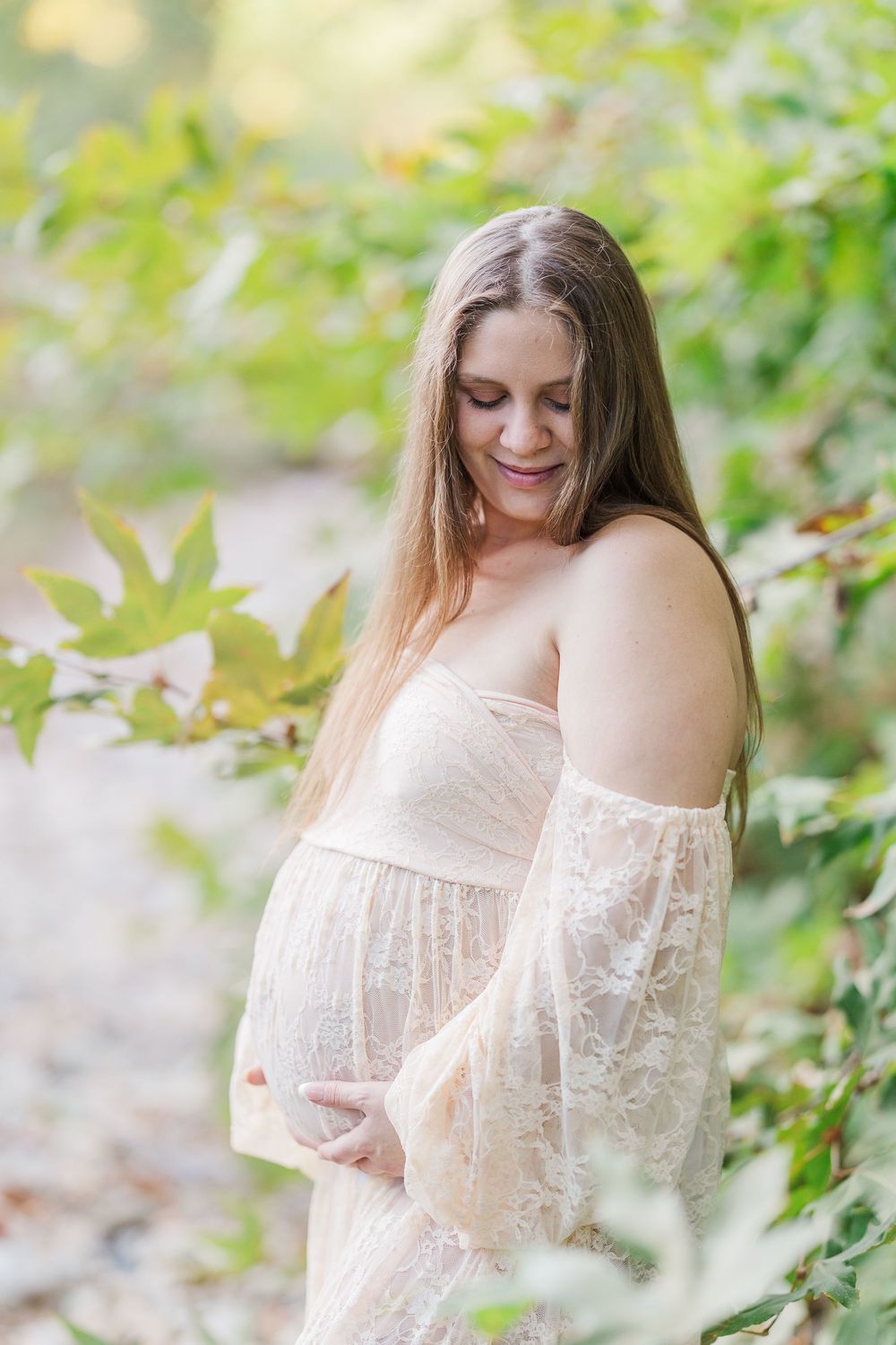 Ethereal maternity photo of an expectant mother in a delicate lace dress posed outdoors among green foliage.