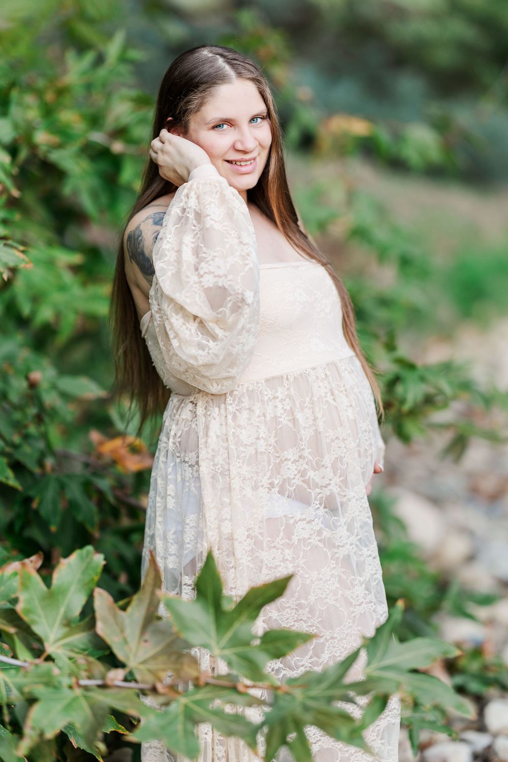 A woman in a delicate lace dress poses gracefully among green foliage in an outdoor setting.