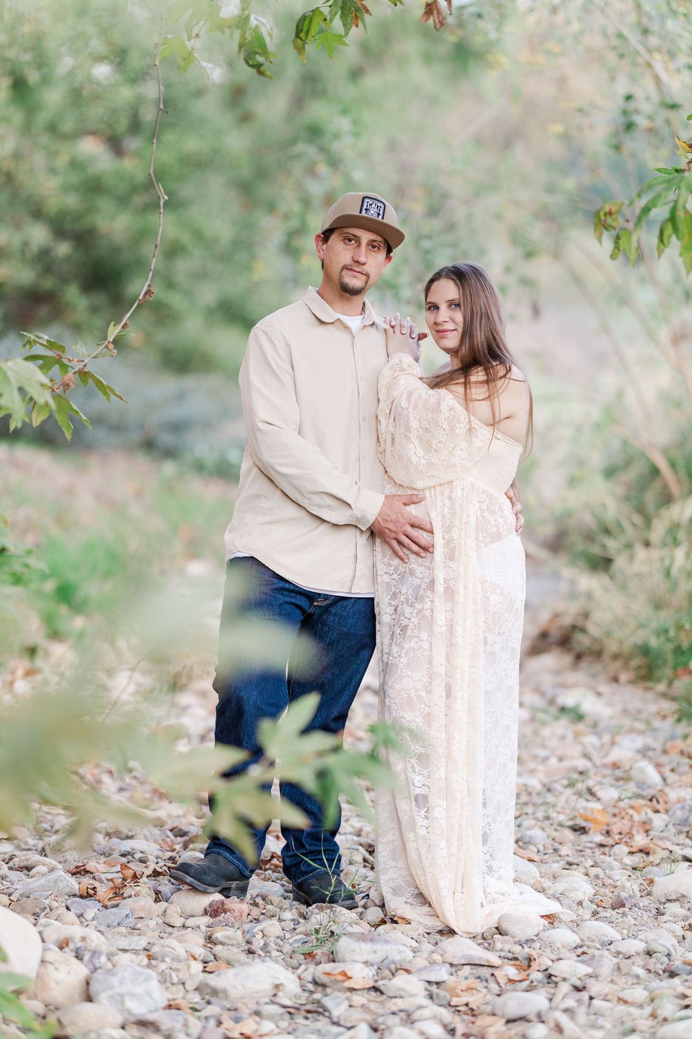 Romantic outdoor maternity portrait of couple in casual attire standing together on rocky ground surrounded by trees.