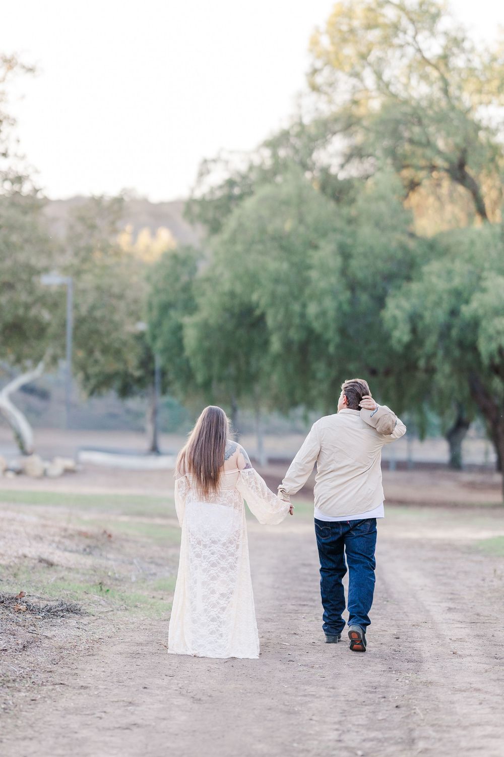 A couple walks hand in hand down a tree-lined dirt path in matching cream-colored outfits.